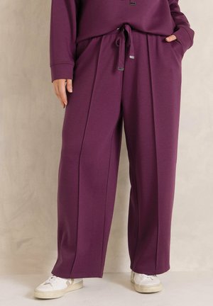 Persoon draagt losse, pruimkleurige broek met een koordsluiting in de taille en witte sneakers, staand met één hand in de zak tegen een neutrale achtergrond.