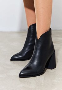 Stivali alla caviglia in pelle nera con design a punta, tacco alto a blocco e un'apertura a V distintiva lungo il gambale. Texture liscia.