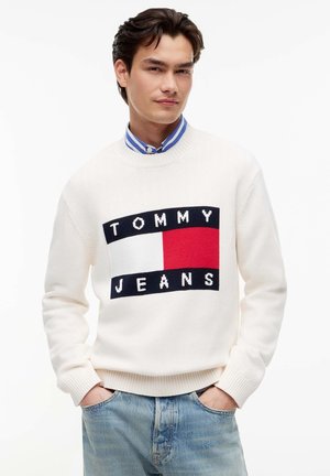 Jeune homme portant un pull blanc Tommy Jeans par-dessus une chemise à col rayée bleue et un jean bleu clair, les mains dans les poches.