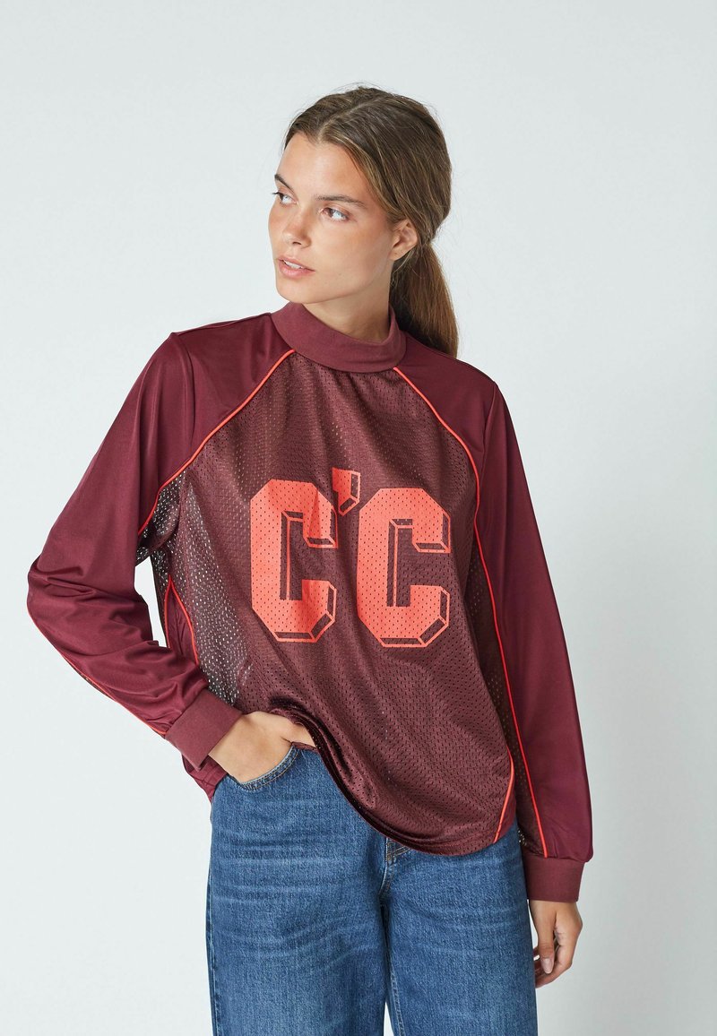 Burgundin pitkähihaisessa paidassa on verkkopaneelit, suuret oranssit "C'C"-printit, ribattu kaulus ja kontrastoivat punaiset yksityiskohdat.