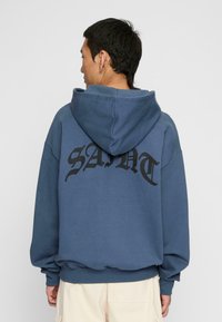 Felpa con cappuccio blu in morbido tessuto, caratterizzata da grande scritta nera "SAINT" sul retro, con vestibilità ampia e polsini a costine.