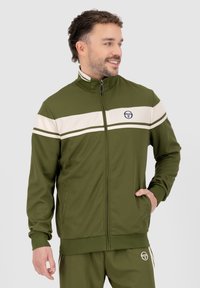 Veste de survêtement vert olive avec des rayures horizontales blanches, fermeture éclair sur le devant, col haut et logo. Fabriquée en tissu lisse, elle est dotée de poches latérales.