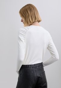 Lyhytukkainen nainen valkoisessa, pitkähihkaisessa ribbineuletussa yläosassa ja mustissa nahkahousuissa, kuvattuna takaapäin vaaleaa taustaa vasten.