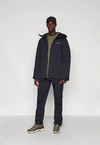 Schwarze wasserdichte Jacke mit Reißverschluss, kombiniert mit schwarzer Hose und Wanderschuhen. Verfügt über eine Kapuze und ein grünes T-Shirt darunter.