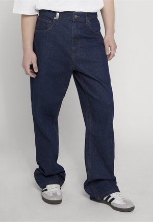 Personne portant un jean bleu foncé ample et des baskets blanches à rayures noires, debout devant un fond clair uni.