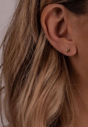 Goldener Ohrring in Llama-Form, der in der Nähe eines Ohrs präsentiert wird, mit polierter Oberfläche und minimalistischen Design, vor glattem, blonden Haar.