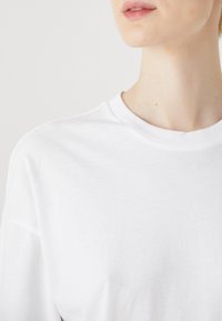 Tricou alb din bumbac, cu guler rotund și mâneci scurte, având o textură netedă și o croială lejeră.
