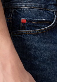 Tmavomodré džíny z denimu majú oranžové šitie a malú červenú nálepku "HUGO" na prednej vrecku. Hladká textúra s jemným opotrebením.