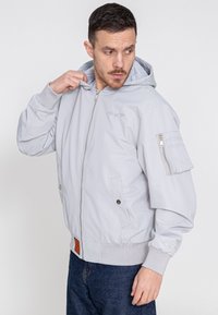 Hellgraue Bomberjacke mit Reißverschluss, elastischen Bündchen, Kapuze und Seitentaschen; verfügt über eine Reißverschlusstasche am Ärmel.