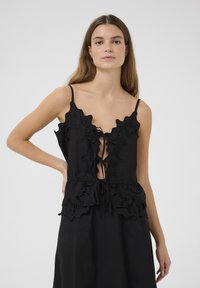 Black top with ruffled edges, adjustable ties at the front, and thin straps. Fabric has a smooth texture, with a relaxed fit silhouette.