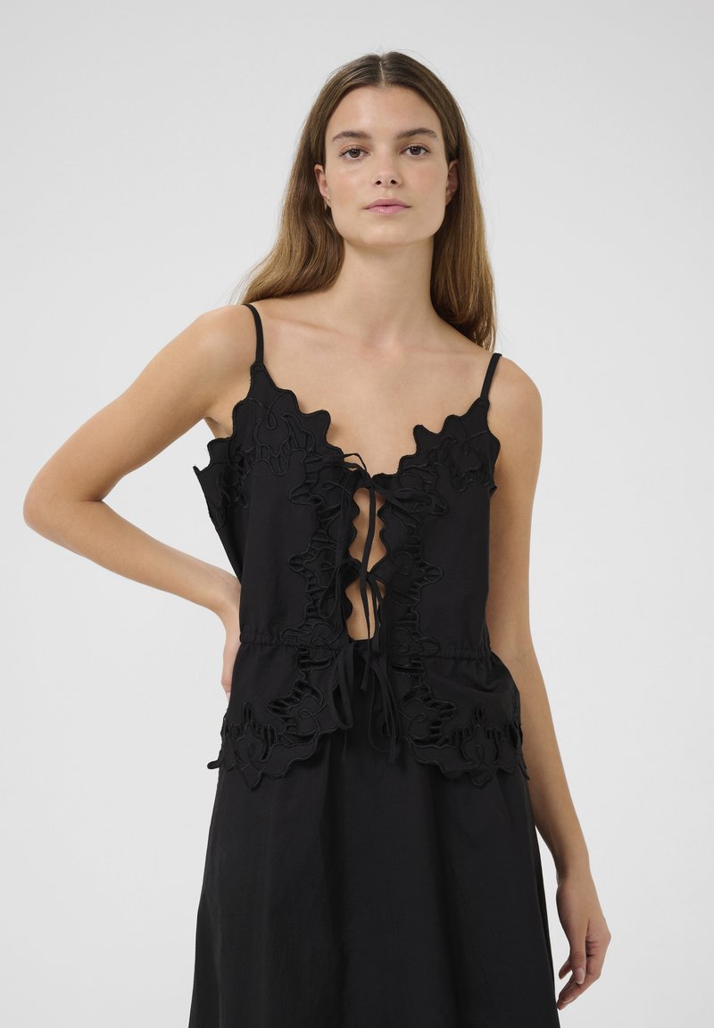 Black top with ruffled edges, adjustable ties at the front, and thin straps. Fabric has a smooth texture, with a relaxed fit silhouette.