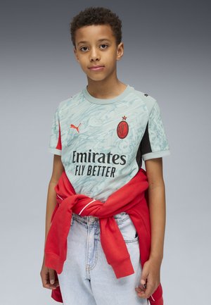 AC Milan Trikot in Hellblau mit strukturiertem Muster, roten Akzenten und schwarzen Ärmeln. Kombiniert mit hochTaillierten Jeansshorts und einem roten Hoodie, der um die Taille gebunden ist.