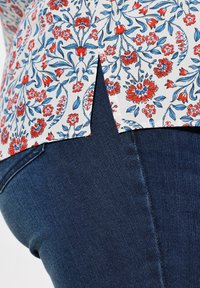 Blusa floral con patrones rojos y azules sobre un fondo blanco, con un dobladillo partido. Combinada con jeans de mezclilla oscuros, que muestran una textura suave.