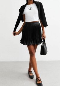 Zwarte geplooide rok, witte cropped T-shirt met "PARIS" graphic, zwarte cardigan over de schouders, zwarte handtas, en zwarte schoenen met gespen.