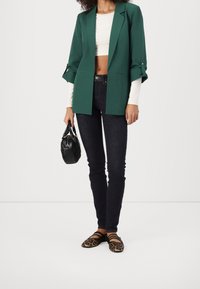 Blazer vert à ouverture frontale, top blanc court en dessous, jean foncé, sac à main noir, et ballerines imprimé léopard. Manches évasées sur le blazer.
