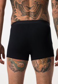 Black cotton boxer briefs with a soft waistband, featuring a snug fit. The background displays intricate colorful tattoos on the skin.