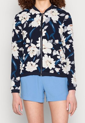 Femme portant une veste zippée bleu marine avec un grand imprimé floral blanc et un short bleu clair sur fond uni.