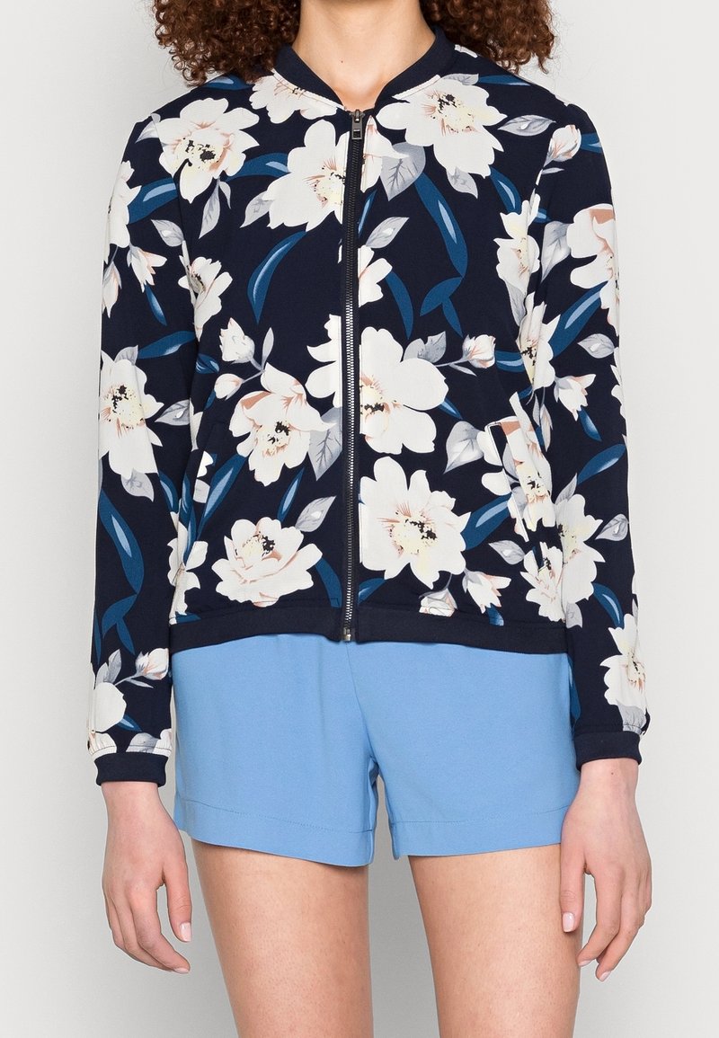 Femme portant une veste zippée bleu marine avec un grand imprimé floral blanc et un short bleu clair sur fond uni.