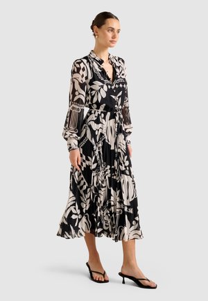Femme debout dans une robe midi à manches longues à motifs noir et blanc avec une jupe plissée et des sandales à talons noires.