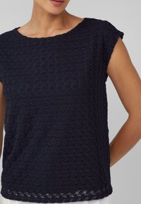 Haut bleu marine avec un motif géométrique texturé, comportant un col rond et des manches courtes. L'ourlet présente un délicat détail en dentelle.