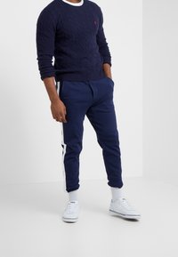 Mörkblå kabelstickad tröja med vit halsringning, kombinerad med mörkblå träningsbyxor med vita sidostråk och vita sneakers.