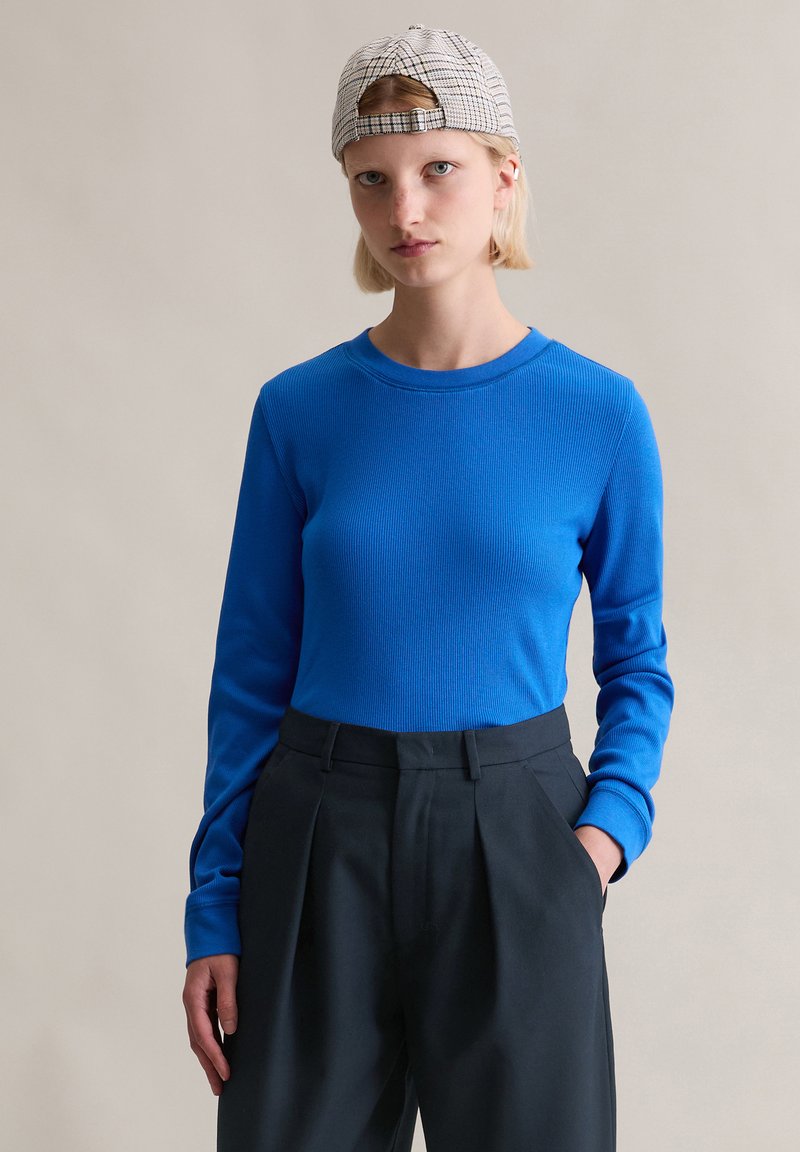 Blaues, geripptes Langarmoberteil kombiniert mit hoch taillierter schwarzer Hose mit Seitentaschen und gemusterter Kappe. Einfaches Design, figurbetonte Silhouette.