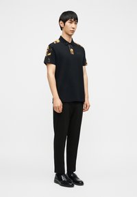 Νέος άνδρας που στέκεται, φοράει μαύρο πόλο με χρυσά σχέδια στους ώμους, μαύρο παντελόνι και μαύρα δερμάτινα loafers, μπροστά σε απλό λευκό φόντο.