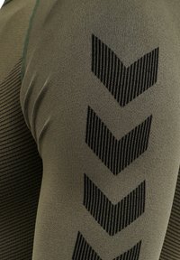 Olivgrön träningströja med strukturerad tyg; har svarta diagonala ränder och chevronmönster längs ärmen.