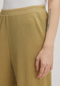 Licht olijfgroene, textuurlijke broek met een elastische tailleband, met een golvende stofafwerking. Een hand rust op de stof, wat de val benadrukt.