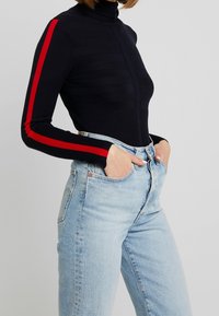 Pull à col roulé bleu marine avec des rayures rouges sur les manches, texture côtelée. Associé à un jean taille haute bleu clair, avec une coupe classique.