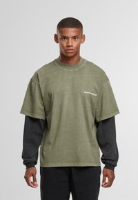 Noor mees lühikeste juustega, kandes kihilist rohelise värvi lühikeste varrukatega T-särki musta pikkade varrukatega särgi peal, vaatab otse, neutraalne taust.