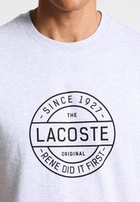 Graues Baumwoll-T-Shirt mit einem kreisförmigen schwarzen Logo und dem Text: "Seit 1927 hat Lacoste Original Rene es zuerst gemacht." Klassisches Rundhalsausschnitt-Design.
