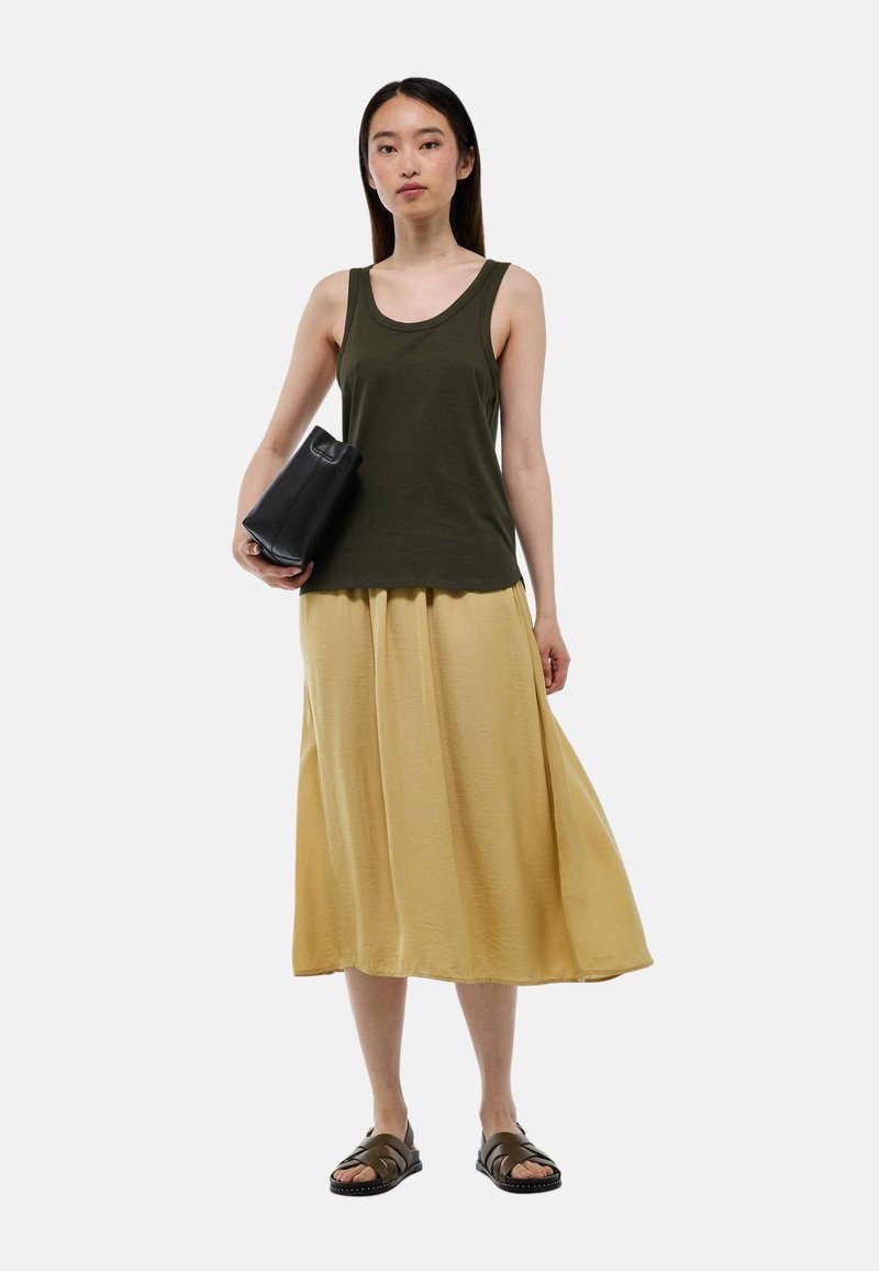 Débardeur vert olive associé à une longue jupe jaune douce. Un sac noir et des sandales foncées complètent la tenue. Design simple et tissu léger.
