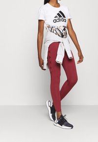 Vit kortärmad Adidas-t-shirt med svart logotyp, röda leggings med svarta sidodetaljer, och svarta träningsskor, i kombination med en grå tröja knuten runt midjan.