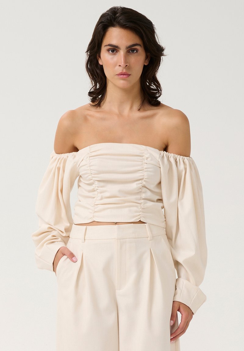 Vállnélküli krém színű crop top, fodros részletekkel, hosszú puff ujjakkal és testhezálló siluettel. Sima anyag texturált felülettel.
