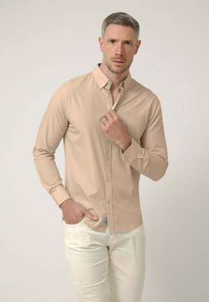 Hombre con cabello gris corto vistiendo una camisa beige con botones y pantalones blanco roto, una mano ajustando el cuello, la otra en el bolsillo.