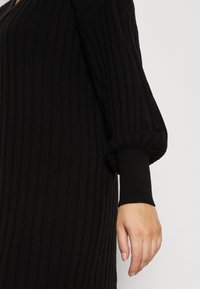 Robe pull en maille noire côtelée avec col en V, manches ballon et poignets ajustés, mettant en valeur la texture et une silhouette élancée.