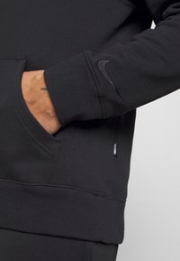 Sweat-shirt Nike en tissu noir, avec une poche avant, des poignets côtelés, et un petit logo noir sur la manche. Texture lisse.