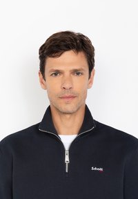 Pullover blu navy con zip realizzato in morbido tessuto, dotato di colletto alto e logo ricamato sul petto. Texture liscia, design semplice.