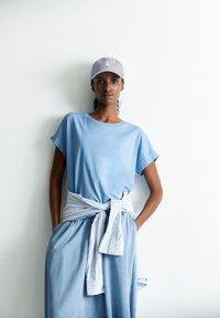 Helles blaues Baumwoll-T-Shirt mit hochgeschlagenen Ärmeln, kombiniert mit einem hellen Jeansrock und einem hell gestreiften Wickel. Graue Kappe mit Logo-Akzent.