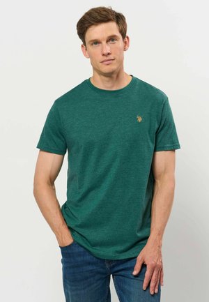 Jeune homme aux cheveux courts et bruns, portant un t-shirt vert à manches courtes et un jean bleu, debout avec une main dans la poche sur un fond uni.