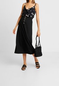 Svart midikjol med knappdetaljer, tillsammans med en blommig camisole. Modellen håller en vävd svart väska och bär svarta sandaler med två remmar.