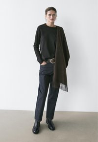 Kvinde iført en sort langærmet skjorte, mørke jeans med bælte, sorte støvler og et frynset mørkebrunt halstørklæde lagt over den ene skulder.