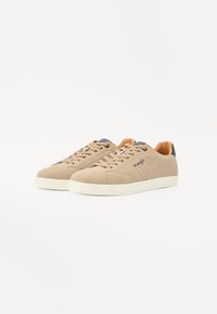 Beige leather sneakers with perforated detailing, round toe, and contrasting dark heel accent. Rubber sole with a textured finish.