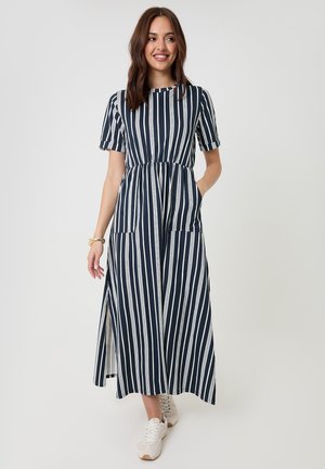 Mujer sonriendo, vistiendo un vestido midi de rayas verticales en azul marino y blanco con mangas cortas y zapatillas blancas, de pie con una mano en el bolsillo.