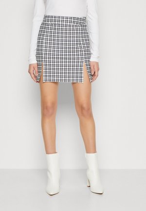 Femme portant une mini-jupe pied-de-poule noire et blanche avec des fentes à l'avant, un haut blanc à manches longues et des bottines blanches, debout devant un fond uni.