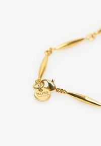 Goldene Kettenarmband mit länglichen, spitz zulaufenden Gliedern. Enthält einen Karabinerverschluss und einen runden Anhänger mit erhabenem Text. Glatte, glänzende Oberfläche.