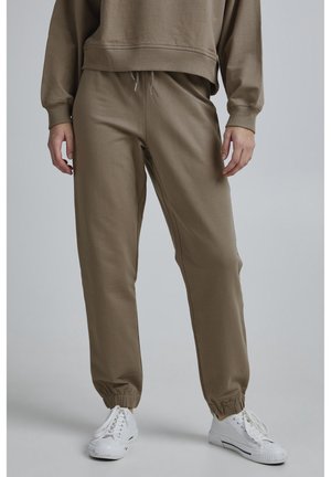 Osoba ubrana w oliwkowozielone spodnie dresowe ze ściągaczami i białe trampki, stojąca na jednolitym jasnym tle.