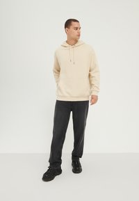 Sudadera beige con cordones ajustables, bolsillo canguro y puños acanalados; combinada con vaqueros oscuros y zapatillas negras.