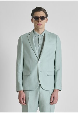 Uomo con abito verde chiaro, polo a coste e occhiali da sole scuri, in piedi davanti a uno sfondo grigio uniforme.